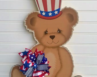 May include: A wooden teddy bear decoration wearing a red, white, and blue Uncle Sam hat. The bear holds a decorative bow with patriotic star and stripe patterns. The bear is hanging on a white slatted surface. The text "JlowsDesigns" is visible.