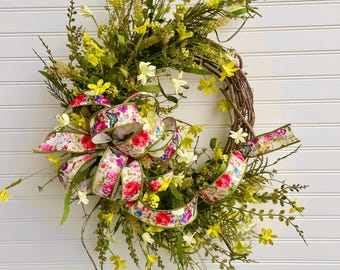 May include: A grapevine wreath adorned with yellow and white wildflowers, and greenery. A large bow with a floral pattern in pink, red, and blue accents the wreath. The text "JlowsDesigns" is visible at the bottom.