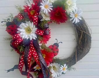 May include: A decorative wreath featuring a grapevine base adorned with red and white daisies, red gerbera daisies, and white daisies. The wreath is embellished with red and black polka dot ribbons and ladybug accents. The overall color scheme is red, white, and black.
