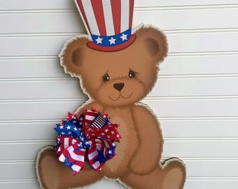 May include: A brown teddy bear-shaped wooden door hanger wearing a red, white, and blue Uncle Sam hat. The bear holds a decorative bow with patriotic colors. The door hanger is suspended by a red cord.