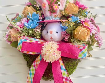 May include: Easter wreath featuring a plush bunny wearing a pink hat with floral accents and bunny ears. The wreath is adorned with colorful flowers, a large pink bow, and a moss base. The bow has a plaid ribbon.