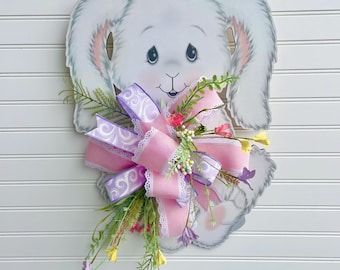 May include: A wooden Easter bunny decoration with a pink and purple bow and floral accents. The bunny is white with gray shading and pink inner ears. The bow is layered with lace and ribbon. The decoration is mounted on a white, horizontal-slat background.