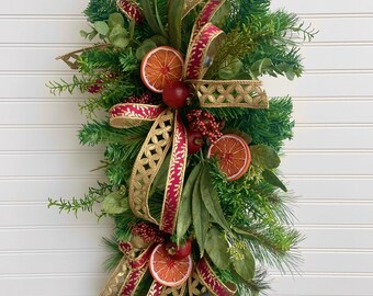 May include: A festive Christmas swag featuring evergreen branches, dried orange slices, red berries, and gold and burgundy ribbons. The swag is adorned with small red apples and blue berries, creating a vibrant holiday decoration.