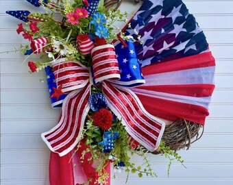 May include: A patriotic wreath with a grapevine base, adorned with red, white, and blue ribbons, fabric, and floral accents. The design incorporates elements of the American flag, including stars and stripes, and is embellished with decorative flowers and greenery.