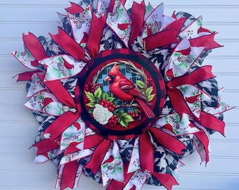 May include: A round wreath with a central image of a red cardinal bird perched on a branch with berries and a white rose. The wreath features red and white patterned ribbons, black and white checkered ribbons, and red ribbons. The wreath is a decorative item.