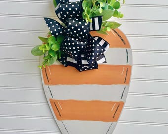 May include: A wooden carrot-shaped door hanger with orange and white stripes. It features a green pumpkin-shaped top, a black and white polka dot bow, and faux greenery. The text "JlowsDesigns" is at the bottom.