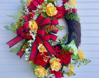 May include: A decorative wreath featuring red and yellow roses, white flowers, and green foliage. A large red bow with black polka dots and ladybug accents add charm. The wreath is displayed against a white background.