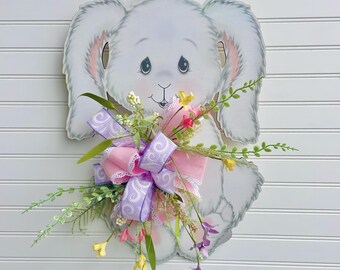 May include: A wooden bunny-shaped wall decoration with a floral arrangement and a decorative bow. The bunny is light gray with pink accents. The bow is pink and purple. The floral arrangement includes yellow and purple flowers.