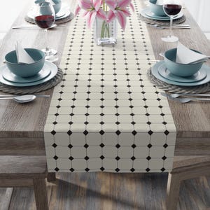 May include: A cream-coloured table runner featuring a repeating black square and diamond design. The runner is laid on a wooden table, set with tableware, wine glasses, and a vase of pink lilies. The table is set for dining.