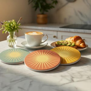 Op de afbeelding: Drie ronde onderzetters met een gestileerd zonnestraalpatroon in gedempt saliegroen, terracotta oranje en goudgeel. Elke onderzetter heeft een witte basis en een glad oppervlak, gerangschikt op een wit marmeren oppervlak bij een koffiekop.