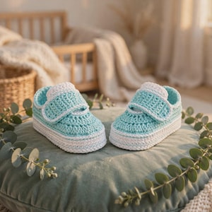 Könnte beinhalten: Ein Paar handgefertigte Babyschuhe aus hellblauem Garn mit weißem Rand. Die Schuhe haben ein strukturiertes Häkelmuster, eine abgerundete Zehenpartie und einen breiten Riemen über dem Spann, der mit einem Klettverschluss befestigt ist.