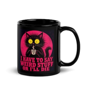 May include: Black ceramic mug featuring a cartoon cat with yellow eyes and a pink background. The cat holds a coffee cup with the words "WEIRD FUEL." The mug has the text "I HAVE TO SAY WEIRD STUFF OR I'LL DIE."