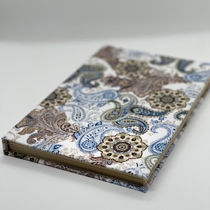 May include: A rectangular journal with a paisley pattern in shades of brown, blue, and cream. The cover features a detailed floral design. The journal is closed and angled, showing the patterned cover and the edge of the pages.