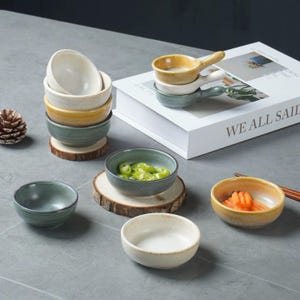 May include: A collection of small ceramic bowls in various colors, including cream, yellow, and green. Some bowls are stacked, while others are filled with food. The bowls are arranged on a gray surface, with a book and a pinecone in the background.
