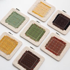 Puede incluir: Un conjunto de posavasos cuadrados con una base de lino natural e inserciones de tela texturizada de colores. Las inserciones presentan varios patrones y colores, incluyendo verde, amarillo y marrón, con bordes deshilachados. Cada posavasos tiene una pequeña etiqueta que dice "LIFE FORMS".
