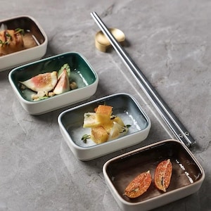 May include: Rectangular ceramic serving dishes with white exteriors and coloured interiors, each containing food. Silver chopsticks and a gold chopstick rest are placed on a grey surface.