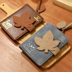 May include: Two wallets, one brown and one denim blue, each featuring a large, tan leaf applique. The brown wallet has gold text and a brown strap. The blue wallet has white text and a tan strap. Both are on a wooden tray.