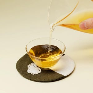 Puede incluir: Un cuenco de vidrio transparente lleno de un líquido dorado, probablemente té o caldo, descansa sobre un posavasos redondo. Una jarra transparente vierte el líquido en el cuenco. El posavasos es una combinación de verde oscuro, blanco y un elemento decorativo de encaje blanco.