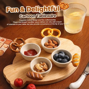 Puede incluir: Una bandeja de madera con cuatro cuencos blancos pequeños llenos de comida. Los cuencos tienen asas con forma de pretzel y pan. Los alimentos incluyen almendras, arándanos y salsa. También hay un vaso de limonada y un cuchillo.
