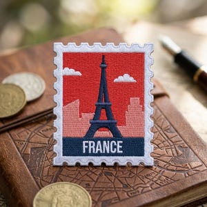 May include: A rectangular embroidered patch shaped like a postage stamp featuring a dark blue Eiffel Tower against a red sky with white clouds and a light pink city skyline. The bottom section is dark blue with the white text FRANCE printed on it.