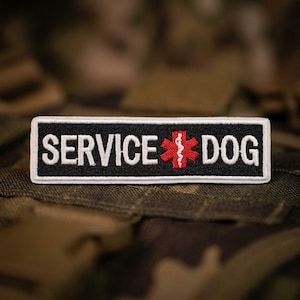May include: A rectangular embroidered patch with a black background and a white border. The text SERVICE DOG is stitched in white capital letters, separated by a red six-pointed star of life symbol featuring a white medical staff and serpent.