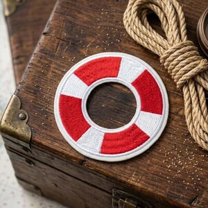 May include: A circular embroidered patch featuring a life preserver design with alternating red and white segments. The patch has a stitched border and a hollow center, showcasing a classic nautical motif with clean, defined lines and vibrant thread.