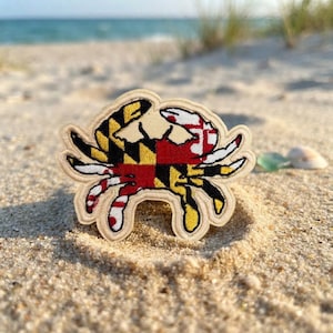 May include: An embroidered patch shaped like a crab, featuring the Maryland state flag design. The crab is divided into four quadrants with black and gold geometric patterns and red and white cross bottony symbols, finished with a tan border.