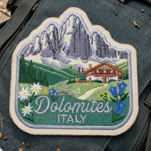 May include: An embroidered patch featuring a mountain landscape with gray peaks, a wooden cabin, green hills, and wildflowers. The text reads Dolomites ITALY in white script and block letters. The patch has a light-colored border and is stitched.
