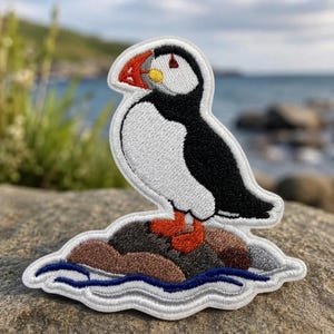 May include: An embroidered patch featuring a puffin with black and white feathers, a bright orange and yellow beak, and orange feet. The bird stands on a brown rock base surrounded by stylized blue water waves, all outlined in white thread.