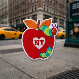 May include: An embroidered patch shaped like a red apple with a brown stem and orange leaves. A green and yellow caterpillar with a red head is on the apple. A white heart in the center contains the red text NY. A second green caterpillar is below.