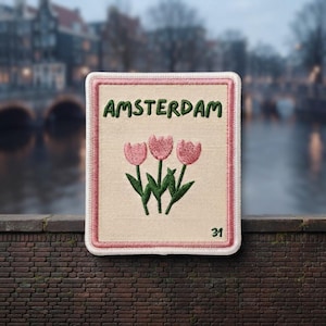 May include: A rectangular embroidered patch with a cream-colored background and a pink border. The text AMSTERDAM is stitched in green at the top. Below, three pink tulips with green stems and leaves are embroidered. The number 31 is in green.
