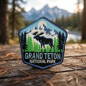 May include: A pentagonal embroidered patch featuring a black moose silhouette standing in front of green pine trees and white snow-capped mountains under a pale blue sky. The text GRAND TETON NATIONAL PARK is stitched in white at the bottom.