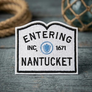 May include: A white embroidered patch with a black border shaped like a sign. It features the black text ENTERING, INC. 1671, and NANTUCKET. A small light blue circular emblem with a central shield design is positioned between the text elements.