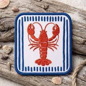 May include: A square embroidered patch featuring a centered orange lobster on a white background, framed by a thick blue border with vertical blue stripes. The patch has rounded corners and a textured fabric surface with detailed stitching.