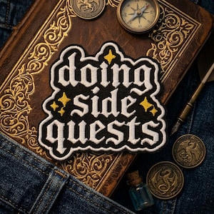 May include: An embroidered patch with a black background and white text that reads "doing side quests" in a gothic font. The patch features small yellow four-pointed star accents and has a scalloped border, showcasing detailed needlework.
