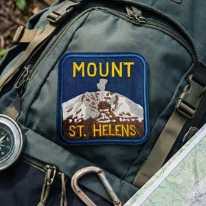 May include: A square embroidered patch featuring a dark blue background with a depiction of Mount St. Helens in white and brown thread. The text MOUNT is in yellow at the top and ST. HELENS is in yellow at the bottom of the patch.
