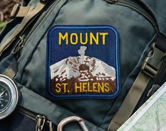 May include: A square embroidered patch featuring a dark blue background with a depiction of Mount St. Helens in white and brown thread. The text MOUNT is in yellow at the top and ST. HELENS is in yellow at the bottom of the patch.