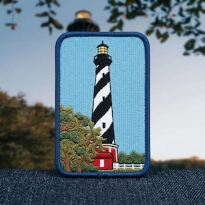 May include: A rectangular embroidered patch featuring a lighthouse with a black and white spiral pattern against a light blue sky. The base of the lighthouse is red, surrounded by green trees and a white fence, all bordered by a dark blue stitched edge.
