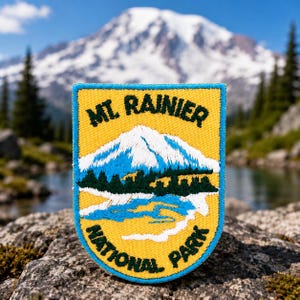 May include: A yellow patch with a blue border featuring an embroidered design of Mount Rainier. The patch includes the text "Mt. Rainier National Park" and a depiction of the mountain with a blue sky and green trees.