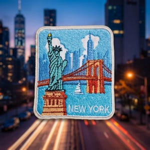 May include: A rectangular embroidered patch featuring a light blue sky background with the Statue of Liberty in teal, a brown Brooklyn Bridge, and a small white sailboat. The text NEW YORK is stitched in white at the bottom of the patch.