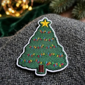 May include: An embroidered patch shaped like a Christmas tree with a green body, a brown trunk, and a yellow star at the top. The tree is decorated with thin, horizontal lines of multi-colored thread representing small festive holiday lights.