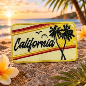 May include: A rectangular embroidered patch with a light yellow background featuring the word California in black script. The design includes two black palm trees, two small black birds, and two horizontal stripes in orange and red.