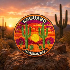 May include: A circular embroidered patch featuring a desert landscape with green saguaro cacti, a bright yellow sun, and orange and purple sky gradients. The text "SAGUARO" is arched at the top and "NATIONAL PARK" is arched at the bottom in white.