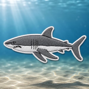 May include: Embroidered patch featuring a gray and white shark with sharp teeth and a dorsal fin. The shark is swimming in a blue ocean with sunlight shining through the water. The patch has a white border.