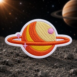 May include: Embroidered patch featuring a stylized planet with orange, yellow, and pink details, surrounded by a white border. The planet has a ring and two pink star accents. The background shows a starry sky and a planet with rings.