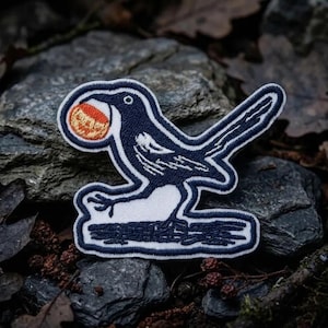 May include: An embroidered patch featuring a dark blue bird with white wing and chest details, perched on a dark branch. The bird holds a small, round, textured orange object in its beak, with a thick white border outlining the entire shape.