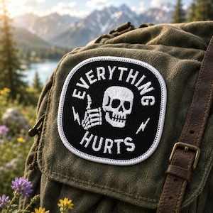 May include: A black and white patch with the words "EVERYTHING HURTS" and a skull with a thumbs-up hand gesture. The patch is sewn onto a green backpack with a brown leather strap. The background features a mountain and lake scene.