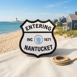May include: A white shield-shaped embroidered patch with a black border. The text reads ENTERING at the top, NANTUCKET at the bottom, and INC 1671 in the middle, flanking a circular blue emblem featuring a shield and an arm with a sword.