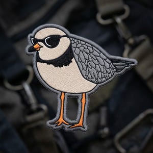 May include: An embroidered patch featuring a bird with light beige plumage, a black neck band, and grey wings. The bird wears black sunglasses and has bright orange legs and a small orange beak, all outlined in a dark grey border.