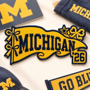May include: A yellow and navy blue embroidered patch with the word "MICHIGAN" in navy blue lettering. The patch has decorative bows and stars. Other items with the same color scheme and text are visible.